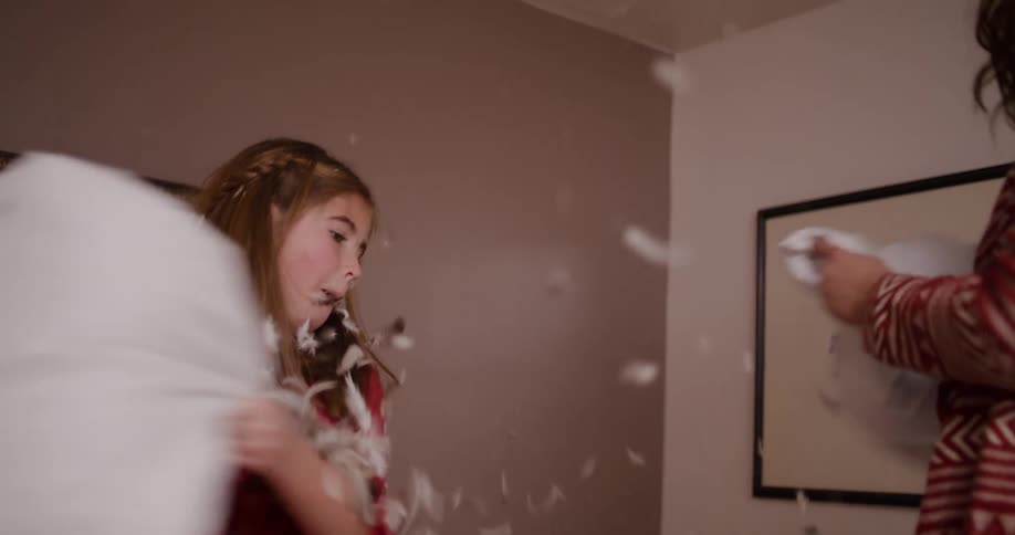 A young girl in a red dress laughs joyfully during a pillow fight as feathers fly through the air in slow motion