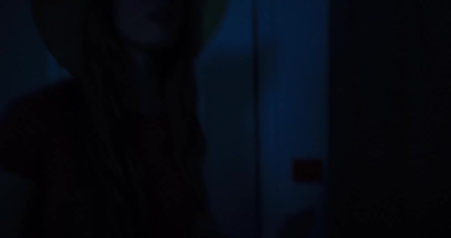 A woman in a sun hat enters a dark hotel room and slowly flips the light switch on
