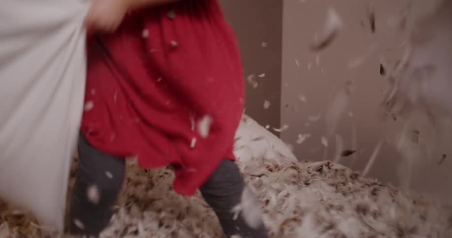 Two happy young girls have an epic pillow fight in slow motion as feathers fly all over the bed