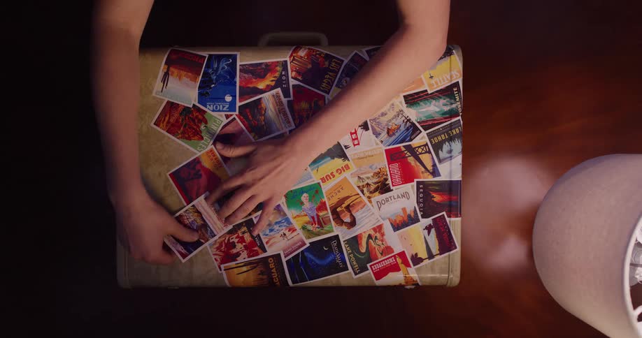From a top-down perspective, a person's hands carefully arrange and stick colorful travel postcards onto a vintage suitcase