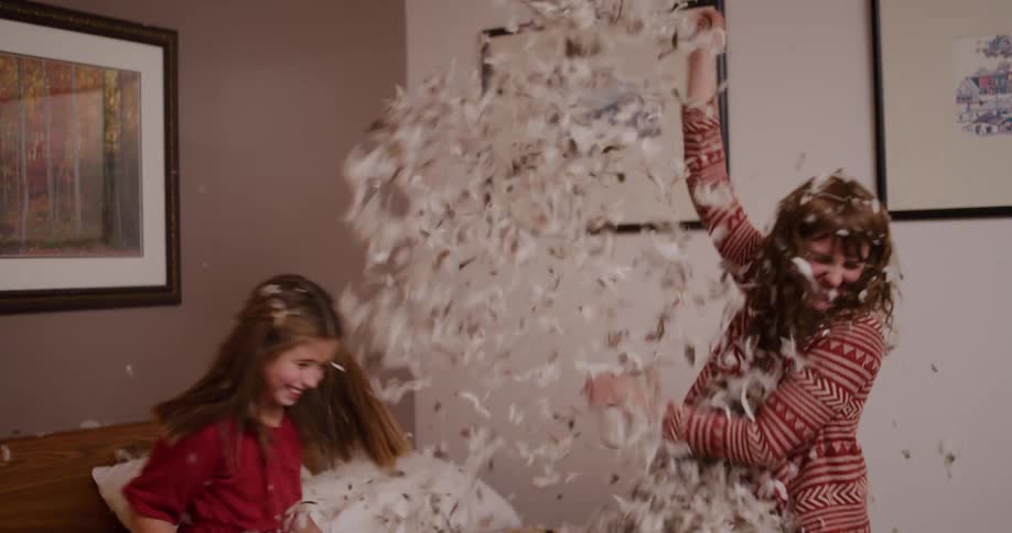 A happy mother and daughter laugh and throw feathers in the air during an epic slow-motion pillow fight on a bed