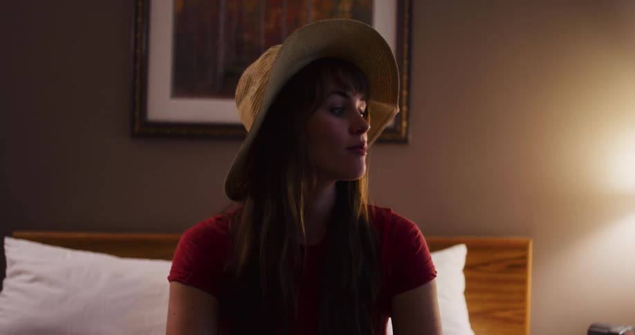 A playful young woman in a sun hat sits on a bed in a hotel room, her mood shifting from energetic to contemplative as the camera pushes in
