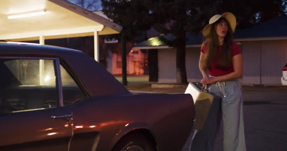 A young woman wearing a wide-brimmed hat takes her luggage out of her vintage car's trunk at a motel parking lot at night