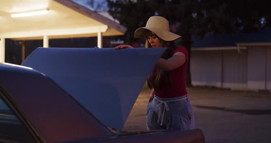 A young woman in a hat slowly pulls a vintage suitcase out of the trunk of her classic car at night