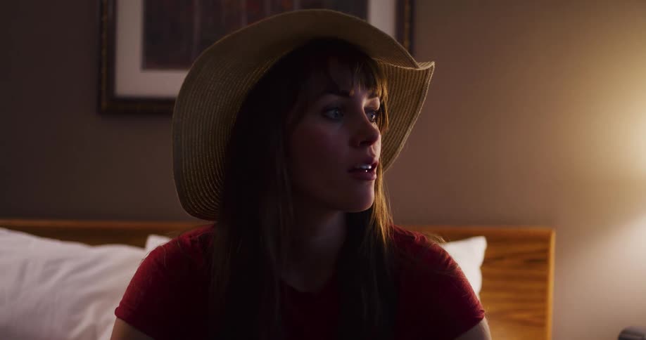 A thoughtful young woman wearing a large straw hat sits on her hotel bed, her expression turning pensive in the dim light