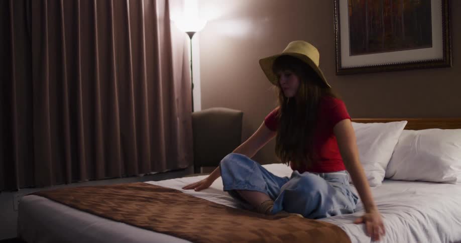 A young woman traveler in a straw hat kicks off her boots and rests on the bed in her dimly lit hotel room