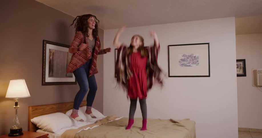 A playful mother and her young daughter laugh while jumping up and down on a bed in slow motion
