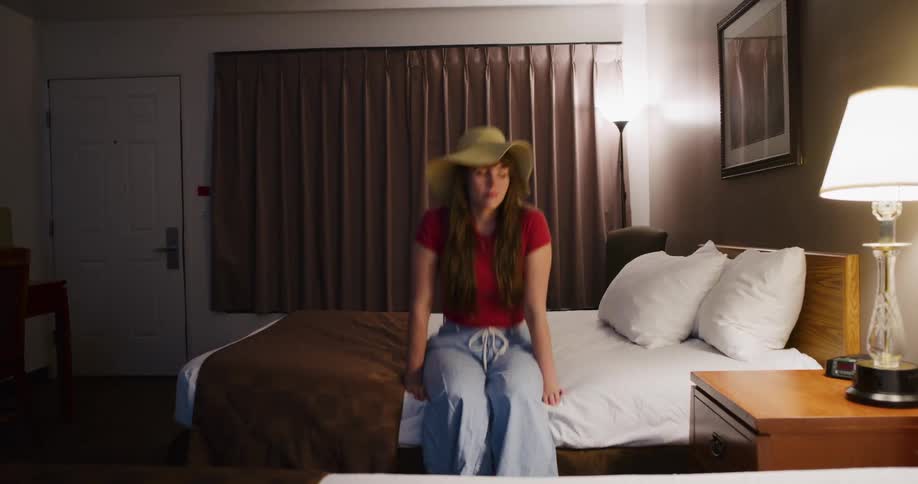 A young woman wearing a large sun hat joyfully jumps and falls back onto her motel bed in slow motion
