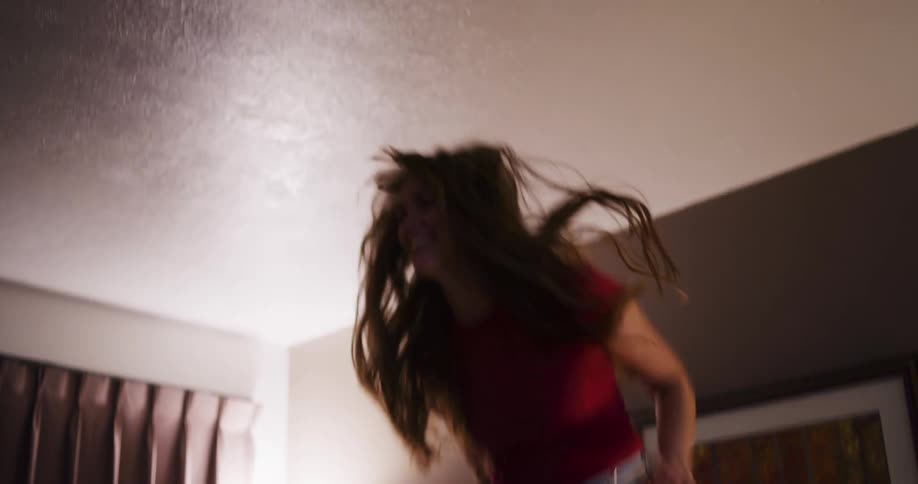 An exuberant young woman in a red shirt and jeans jumps on the bed in slow motion until she happily collapses