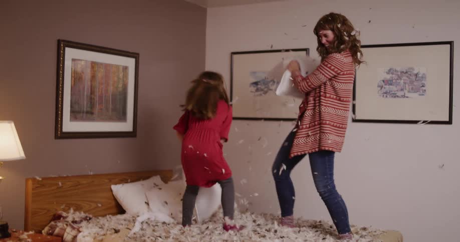 A mother and her young daughter have an epic pillow fight in slow motion, sending feathers flying everywhere as they laugh and jump on the bed