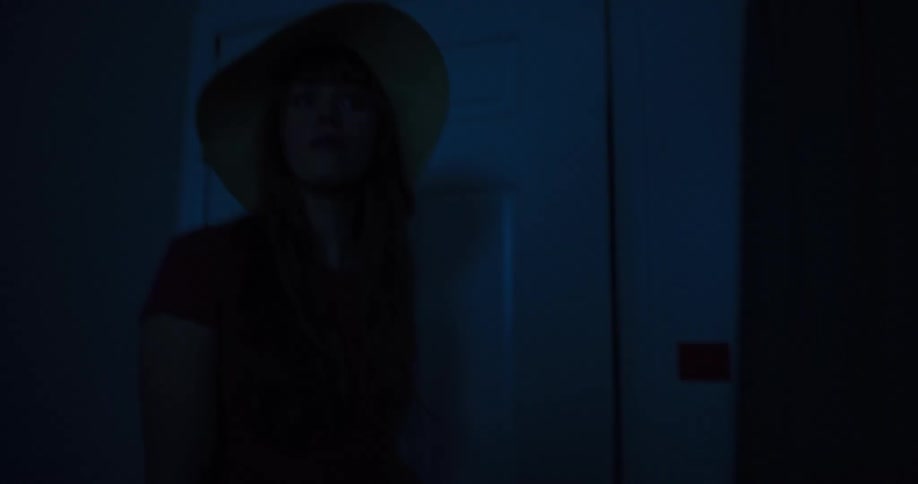A woman cautiously enters a dark room and turns on the light switch, illuminating the space