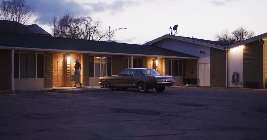 A woman traveler with a vintage suitcase arrives at a roadside motel at dusk, parking her classic car outside her room