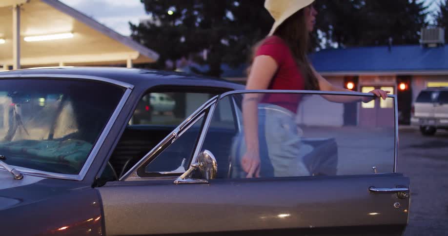 A stylish woman in a hat gets out of her classic grey Mustang in a parking lot at dusk