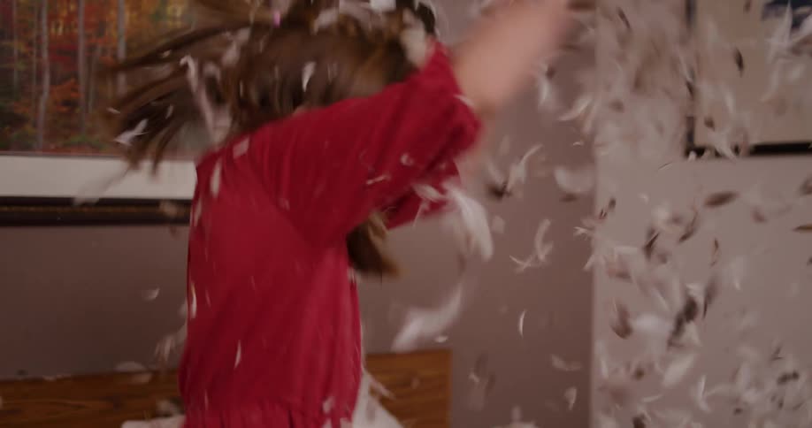 A happy young girl and a woman laugh and play during a chaotic pillow fight as feathers fly everywhere in slow motion
