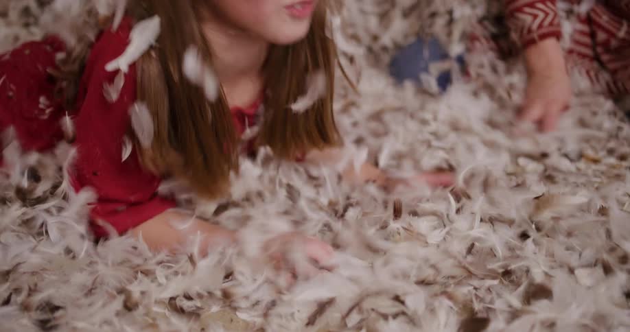 A smiling young girl in pajamas blows on a huge pile of feathers while playing after a pillow fight in slow motion