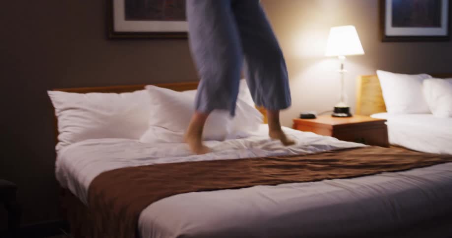 A joyful woman wearing a hat has fun jumping and dancing on a hotel bed in slow motion