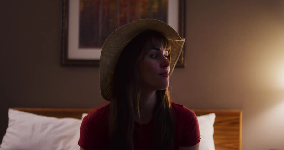 A thoughtful young woman wearing a large sun hat relaxes on a bed in a dimly lit hotel room