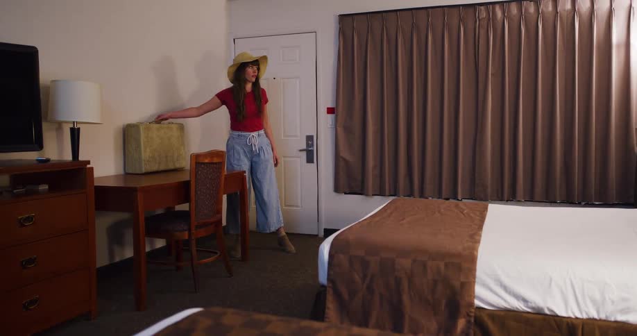 A woman traveler wearing a hat turns on the lights as she arrives in her simple motel room with a vintage suitcase