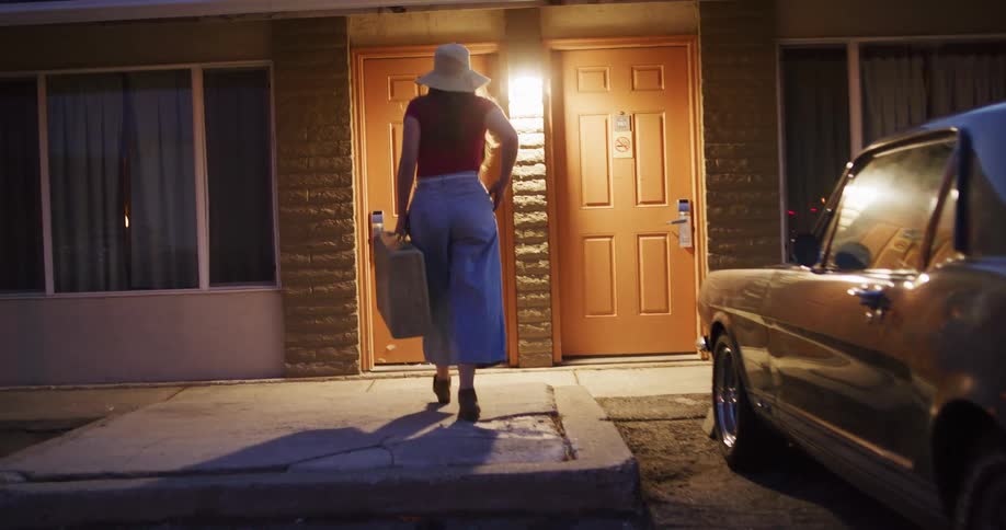 A woman with a vintage suitcase walks from her classic car to her motel room door at dusk