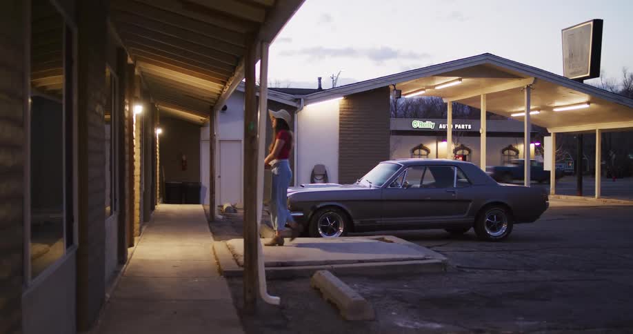 A stylish woman unloads her suitcase from a classic Ford Mustang and walks to her room at a roadside motel at dusk