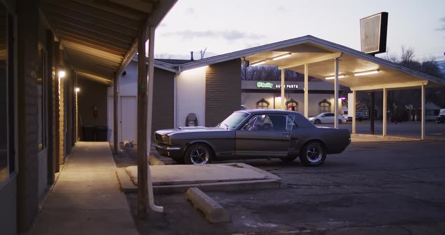 A woman wearing a cowboy hat gets out of a classic Ford Mustang parked at a roadside motel at dusk