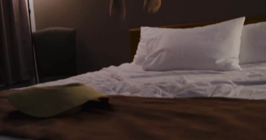 A carefree young woman in a hat excitedly jumps up and down on her motel bed in slow motion before collapsing in laughter
