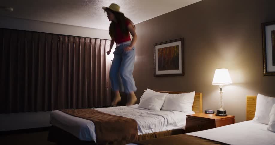 A young woman wearing a hat joyfully jumps up and down on a hotel bed in super slow motion