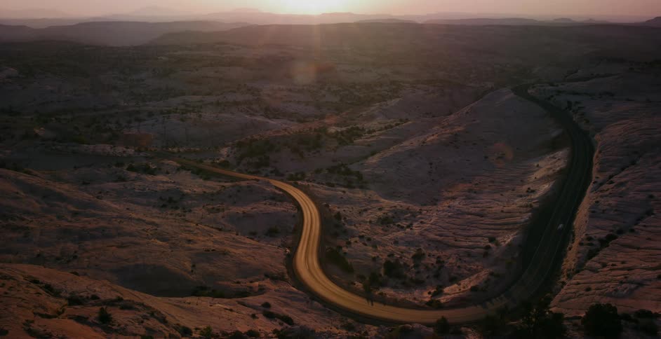 Cars travel down a serpentine highway winding through the desert hills during a golden sunset