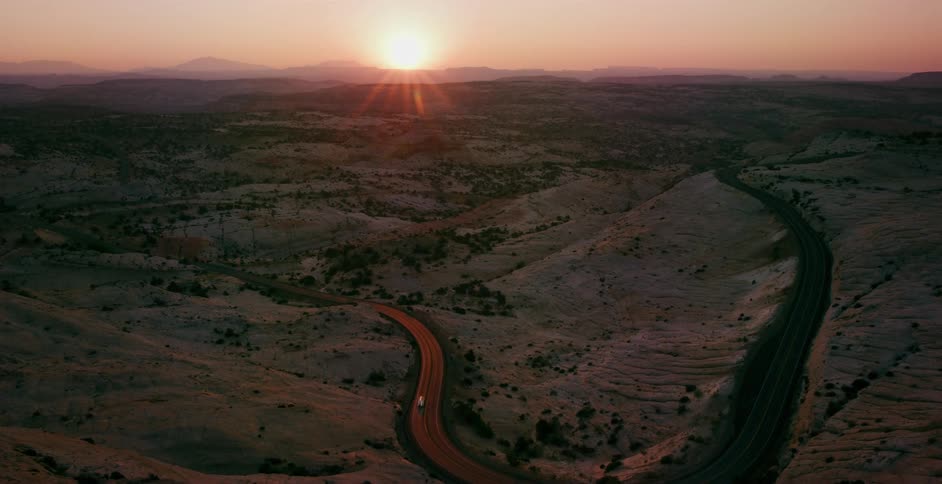 A white truck travels a winding highway through a vast desert landscape as the sun sets beautifully over the distant mountains