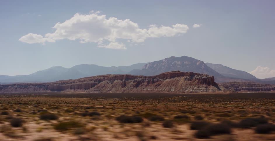 A scenic side-view driving shot captures the vast, arid desert landscape with prominent red rock mesas and mountains in the distance