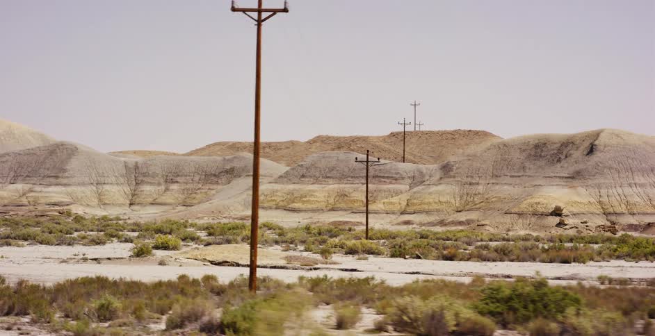 A slow pan across a desolate desert landscape reveals layered badlands and a line of utility poles under a hazy sky