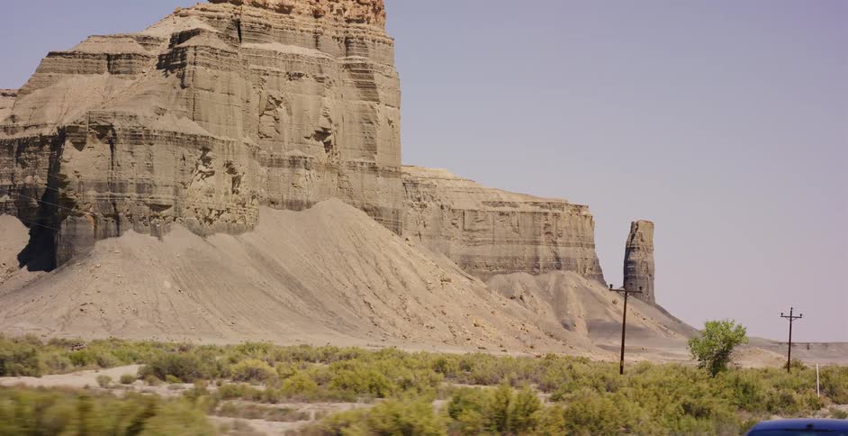 A slow driving shot passes majestic rock formations and desert brush under a clear sky