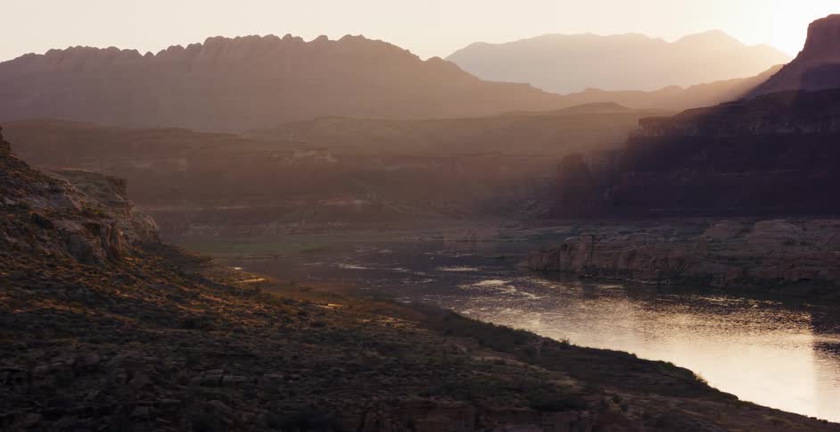 A slow aerial journey over a vast desert canyon as the sun rises, casting a golden glow on the tranquil lake below