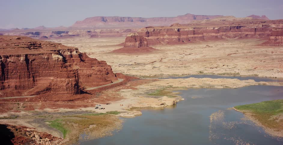 An expansive aerial view reveals a winding lake cutting through a vast, arid red rock canyon landscape on a sunny day
