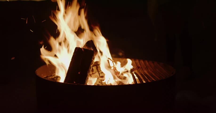 A warm campfire crackles and burns in a metal fire pit in the dark of night