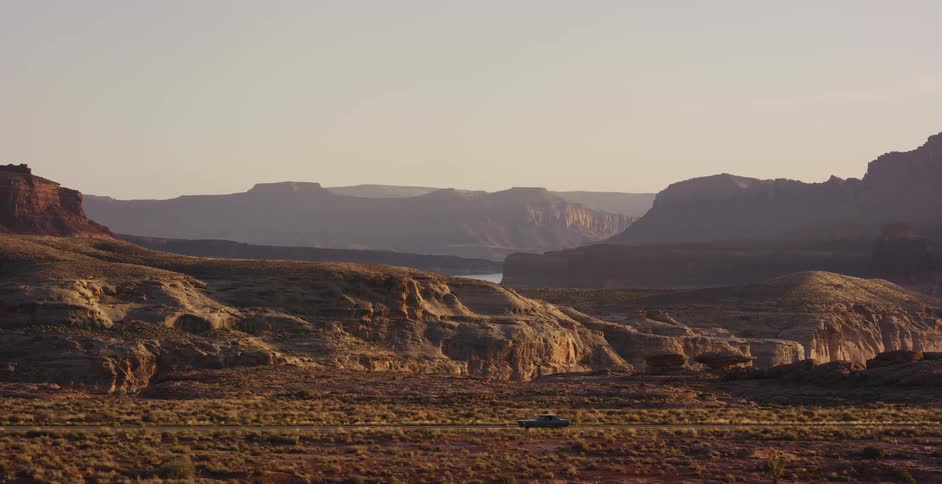 A single car drives down a lonely highway through a vast and beautiful desert canyon landscape during a warm sunset