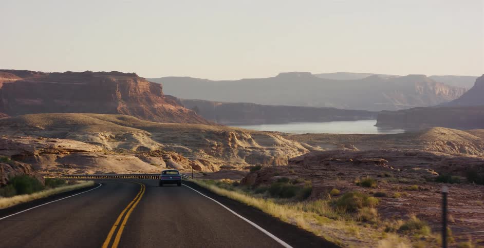 A classic car travels down a winding desert highway, revealing a stunning view of a canyon lake at sunrise
