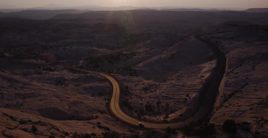 Cars travel along a winding scenic byway through a vast desert canyon landscape as the sun sets
