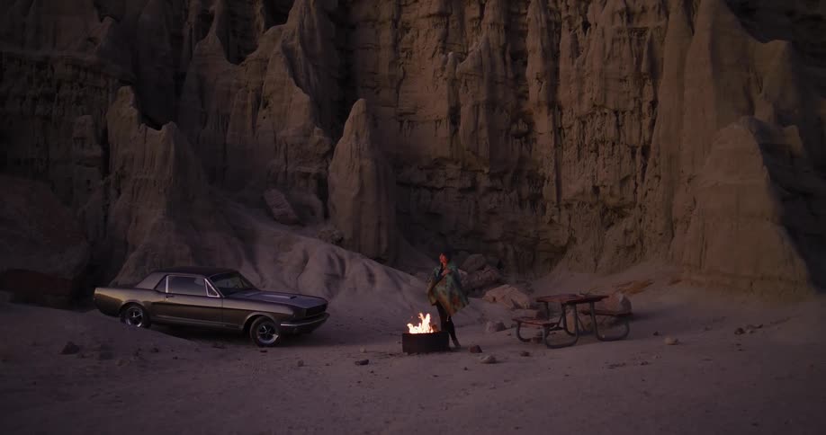 A person wrapped in a blanket stands by a roaring campfire in a remote desert canyon with a classic car at dusk