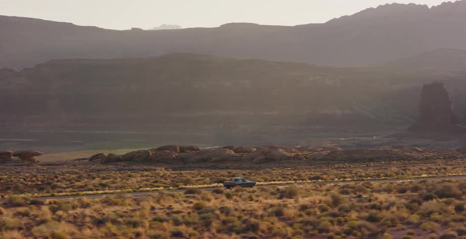 A classic muscle car cruises down a desert highway through a vast canyon landscape at sunset