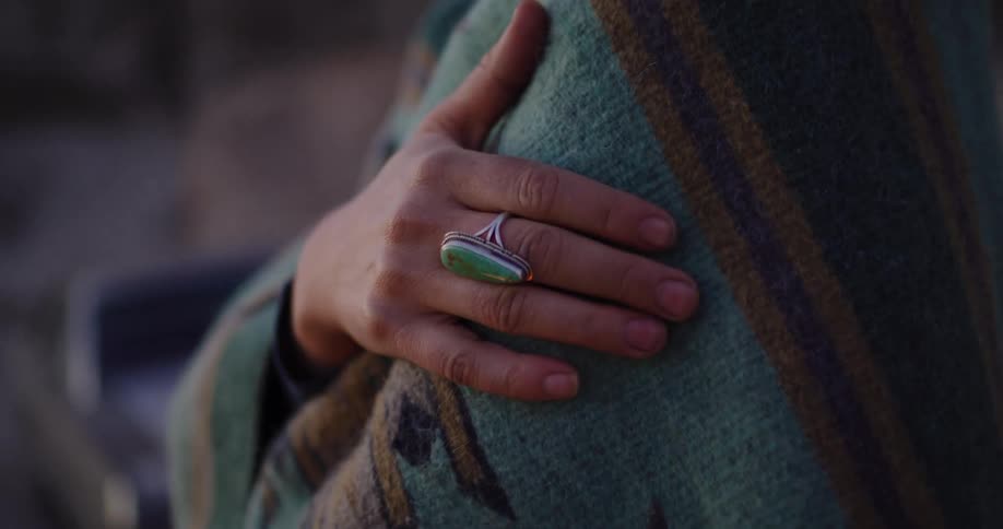 A person with a turquoise ring wraps themselves in a warm, patterned blanket outdoors at dusk