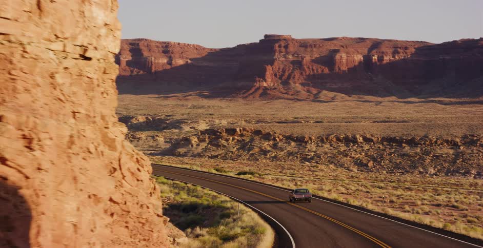 A lone car travels down a winding highway through a vast red rock desert canyon landscape during the golden hour