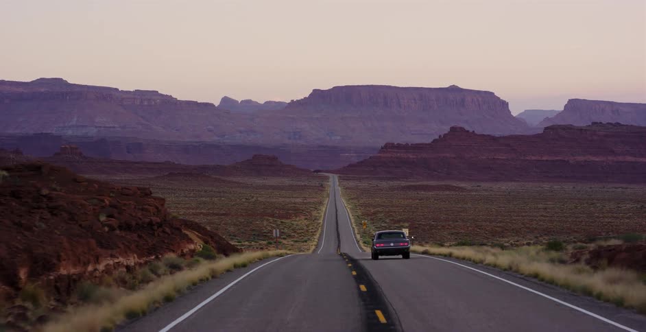A lone car drives down a seemingly endless desert highway toward distant red rock mountains at twilight