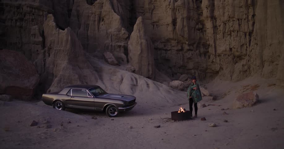 A wide shot shows a person standing by a campfire next to a classic Ford Mustang in a desert canyon