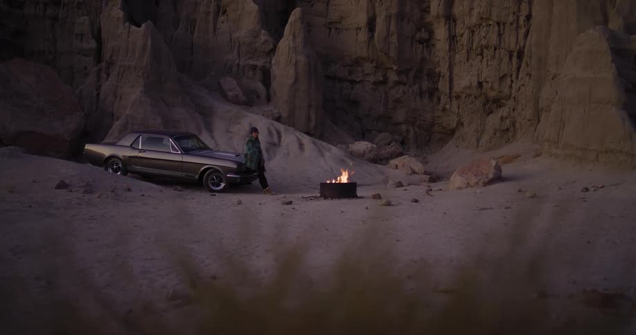 A solitary figure leans against a vintage muscle car next to a campfire in a remote desert canyon at dusk