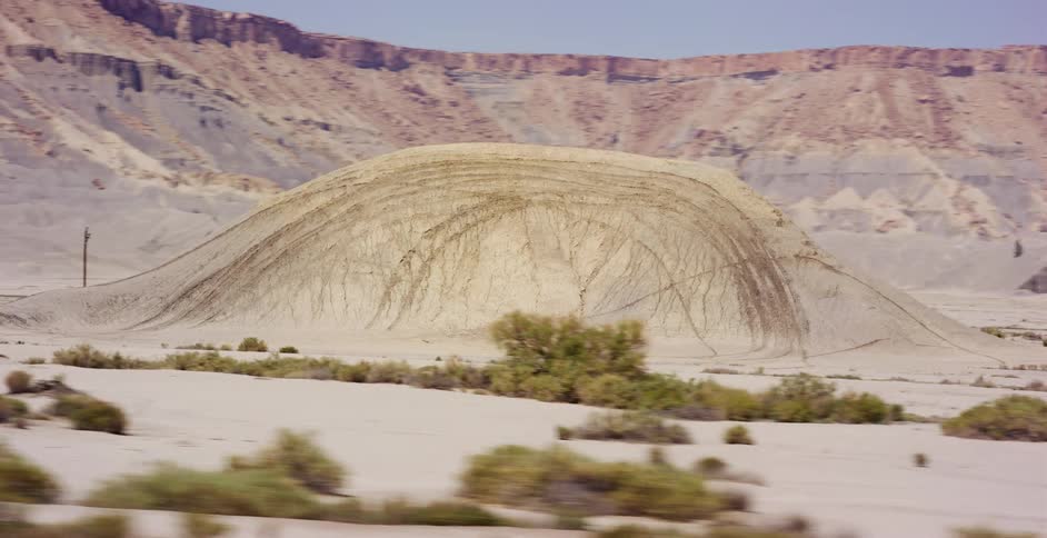 A slow-motion tracking shot from a moving vehicle captures a unique layered rock formation in a vast, arid desert landscape