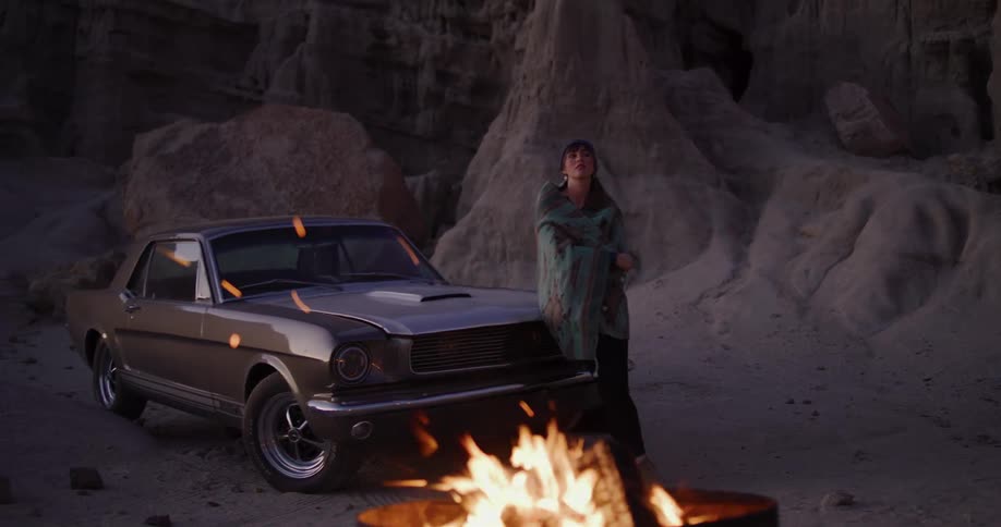 A woman wrapped in a blanket stands next to her classic car by a bonfire in a desert canyon as sparks fly in slow motion