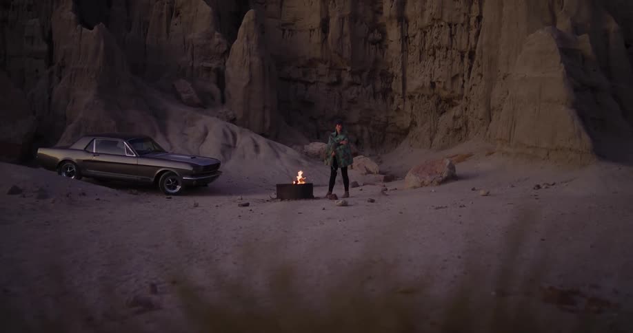 A solitary person stands by a campfire next to a vintage car in the vast, rocky desert badlands