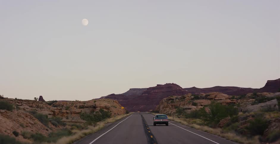 A vintage car drives along a desolate desert highway at dusk, with red rock formations in the distance and the moon overhead
