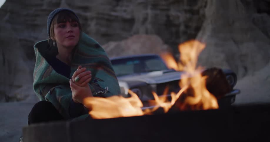 A woman in a beanie finds warmth from a campfire on a cool desert evening during a road trip